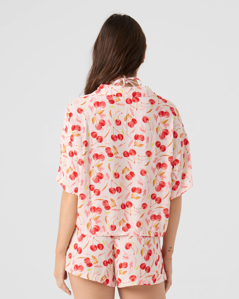 A woman with long brown hair is seen from the back, wearing the Roxy Cherry On Top Crop Stoke Short set with a pink and red cherry print in a light-colored studio.