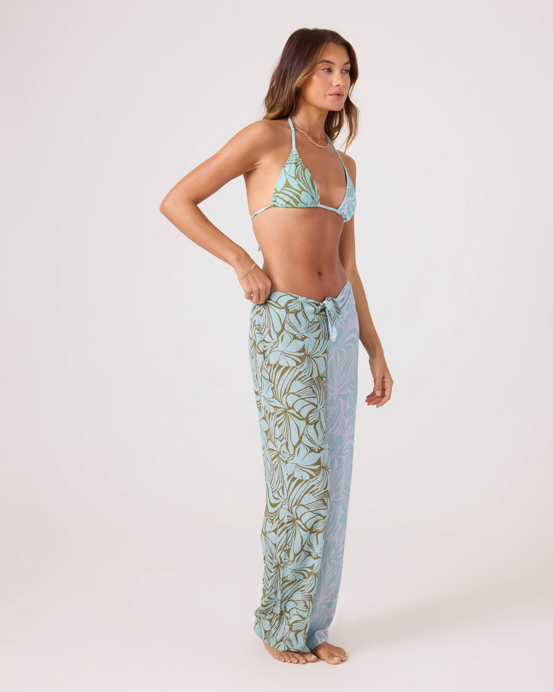 A woman stands barefoot against a plain backdrop, wearing a light blue bikini top and Roxy's Sea Bloom Front Tie Pant—a beach cover-up with an elastic waist and loose, high-waisted blue and green leaf-print fabric. She faces slightly to the side.