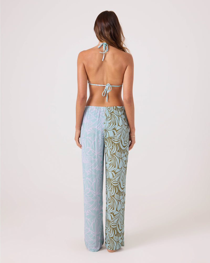 A woman faces away, wearing a halter bikini top and the Roxy Sea Bloom Front Tie Pant—a high-waisted, wide-leg style with elastic waist and mixed pastel and leaf prints—ideal as a chic beach cover-up against a plain light background.