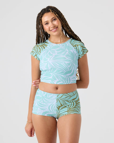 A woman with long braided hair smiles, wearing the Roxy Sea Bloom Crop Rashguard and high-waisted shorts, both in a blue and green leaf pattern, made from Lycra stretch surfwear, standing against a plain light background.