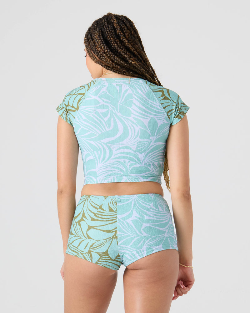 A person with long braids is seen from the back in a matching Roxy Sea Bloom Boy Short and crop top in light blue and green tropical print, made from eco-friendly recycled nylon, against a plain light gray background.