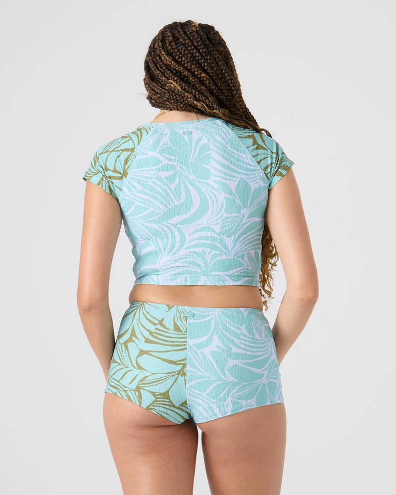 A woman with long braids, seen from the back against a plain light background, wears the Roxy Sea Bloom Boy Short recycled-nylon bikini bottom and matching leaf-print crop top.