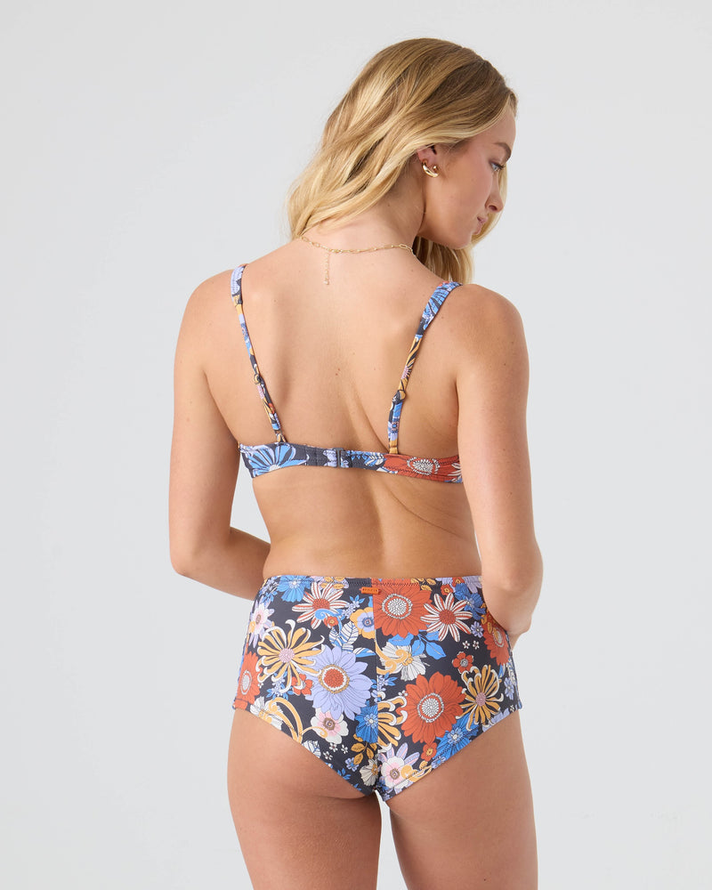 A woman with long blonde hair is seen from the back wearing the Roxy Soul Flower Scoop Bralette Bikini Top, featuring a blue, orange, and yellow floral pattern with adjustable straps, set against a plain light gray background.