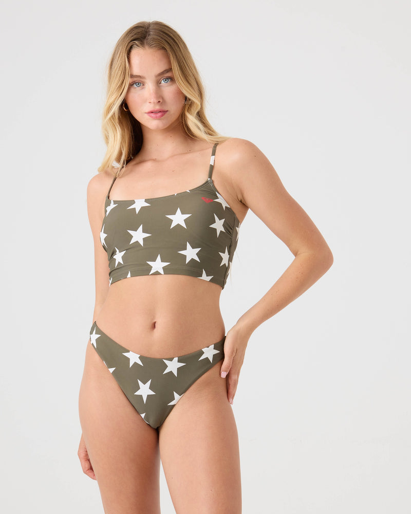 A woman poses in the Roxy Star Daze Scoop Moderate Bikini Bottom, featuring a green and white star print and made from recycled nylon, paired with a matching crop top bikini top against a plain light background.