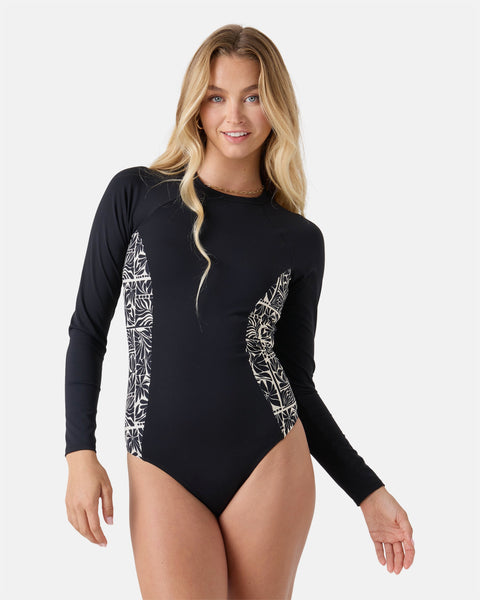 A woman with long blonde hair smiles while wearing the Roxy Geo Flower Surf Suit, a black long sleeve suit with white floral patterned side panels, standing against a plain white background.