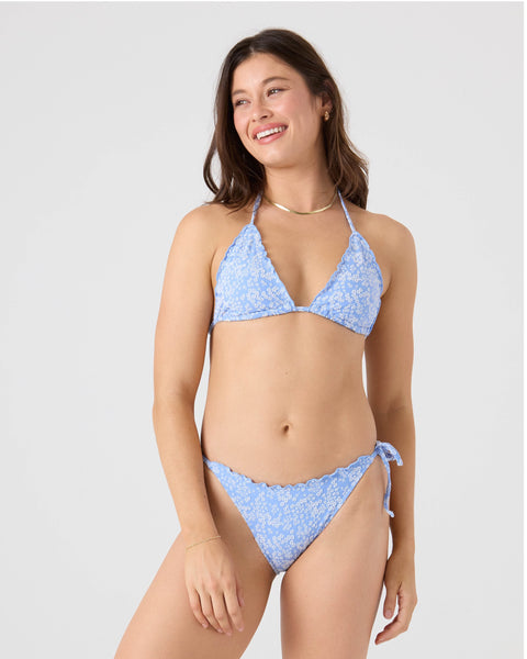 A woman smiles while wearing the Roxy Margarita Tie Side Cheeky Bikini Bottom in light blue floral with scalloped edges, standing against a plain light background.