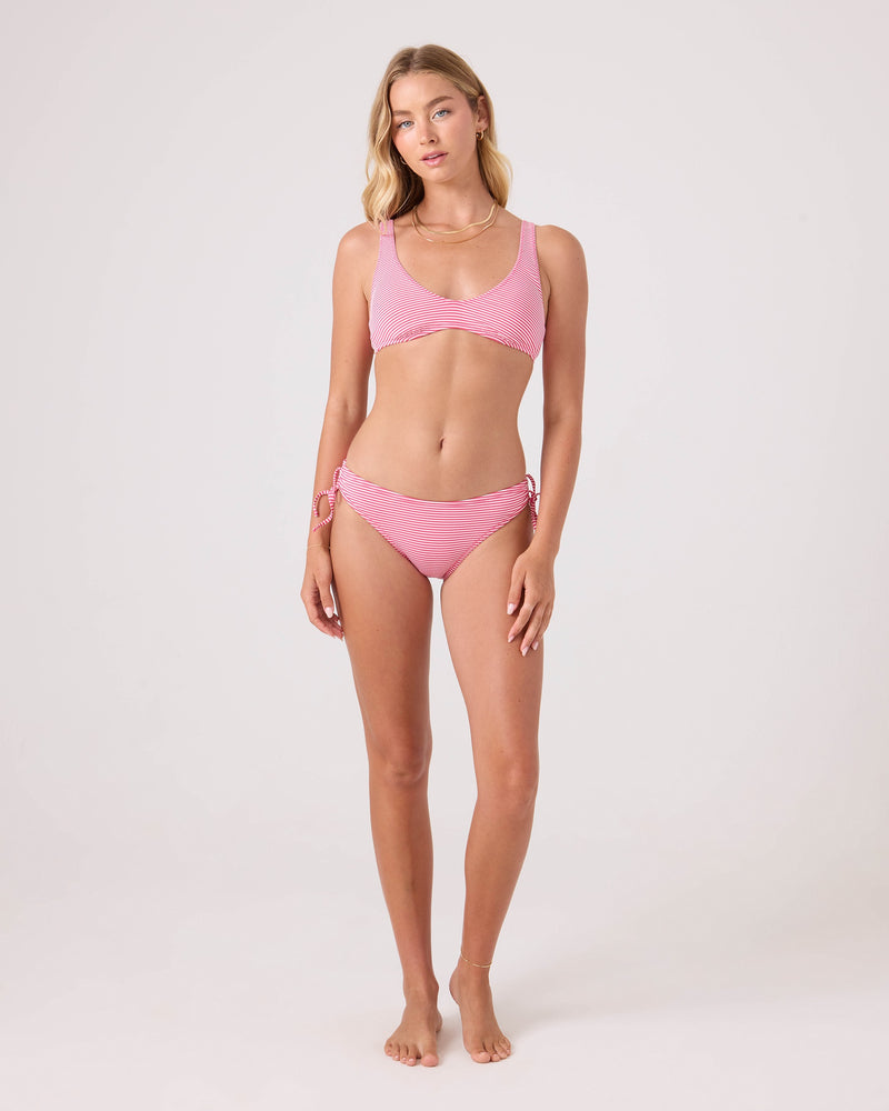A woman with long, wavy blonde hair and a neutral expression stands barefoot on a plain background, wearing the Roxy Good Vibes Tie Back Bralette Bikini Top in light pink with matching side-tie bottoms.