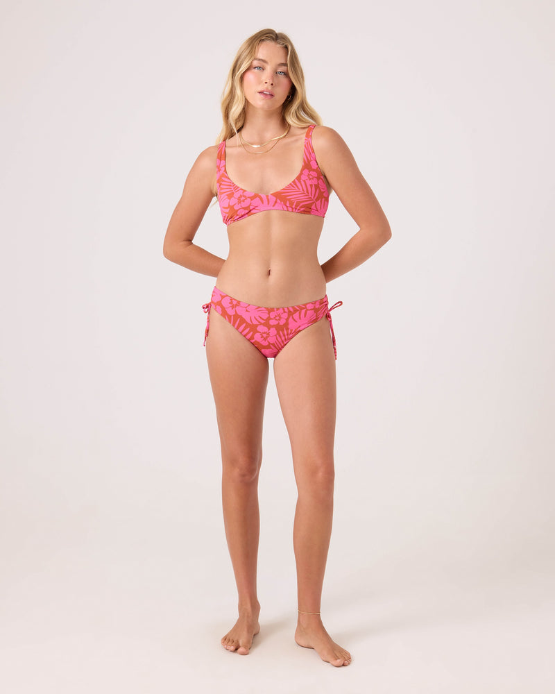 A woman with long blonde hair stands barefoot against a light background, wearing the Roxy Sun Chasers Hipster Lace Full Bikini Bottom in a pink leaf pattern made from recycled nylon, looking relaxed and confident.