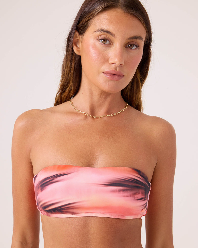 A woman with long brown hair models the Roxy Palm Before The Storm Lace Up Bandeau Bikini Top in pink and black tie-dye, styled with a gold chain necklace against a simple, light-colored background.