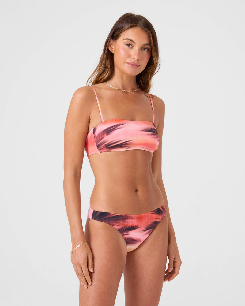 A woman with long brown hair wears the Roxy Palm Before The Storm Hipster Moderate Bikini Bottom, featuring a pink and black print with thin straps, standing against a plain light gray background and looking at the camera with a slight smile.