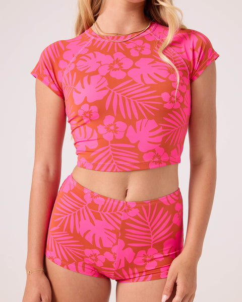 A woman wears the Roxy Sun Chasers Crop Rashguard in Pink Combo with matching high-waisted shorts, both featuring tropical floral and leaf prints. Her face is partially out of the frame.