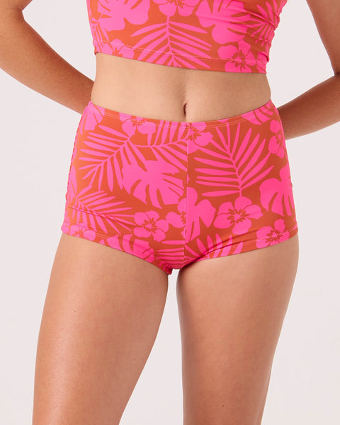 A person wears a Roxy Sun Chasers Boy Short Cheeky Bikini Bottom in Pink Combo with a tropical floral pattern, paired with a cropped top. The background is plain and light-colored.