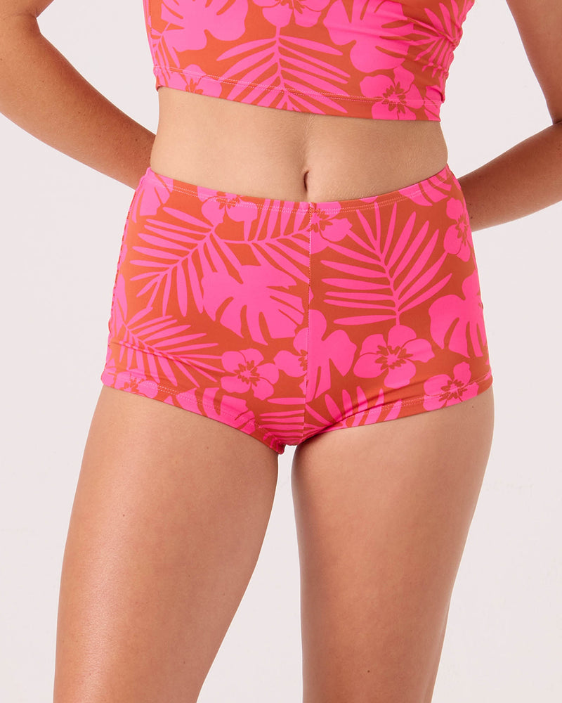 A person wears a Roxy Sun Chasers Boy Short Cheeky Bikini Bottom in Pink Combo with a tropical floral pattern, paired with a cropped top. The background is plain and light-colored.