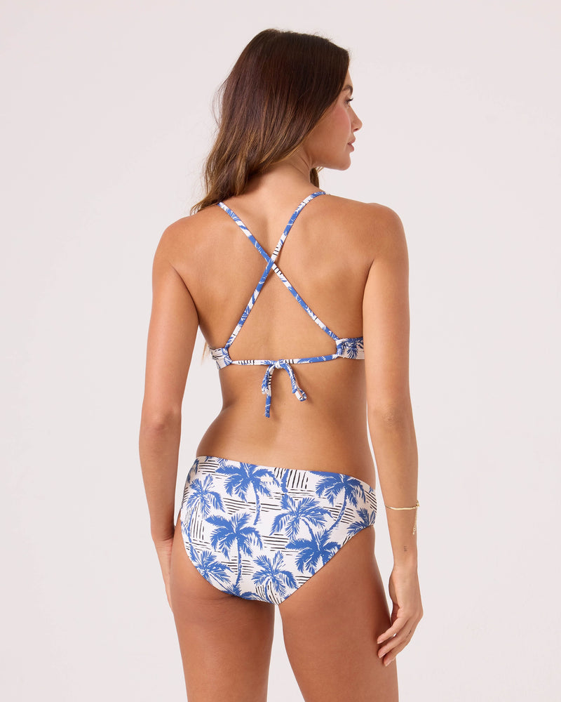 A woman with long brown hair and light skin wears the Roxy Breezy Does It Athletic Triangle Bikini Top, featuring a blue and white palm tree print, crisscross back straps, and a tie-back, standing against a plain background.