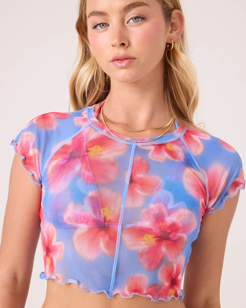 A young woman with long blonde hair wears Roxy's Hibiscus Daze Mesh Crop Top, featuring a blue background with large pink hibiscus prints. She pairs it with gold hoop earrings and layered gold necklaces.