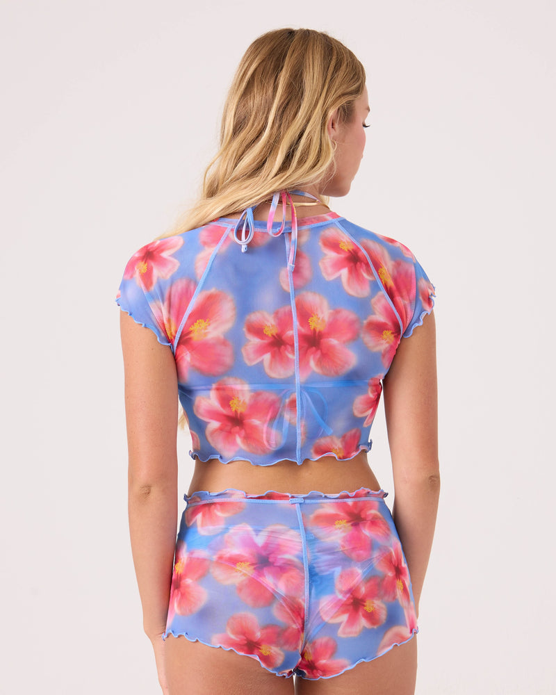 A woman with long blonde hair is seen from the back wearing the Roxy Hibiscus Daze Mesh Crop Top, a sheer blue piece with bright pink and red hibiscus flowers, paired with matching high-waisted shorts.