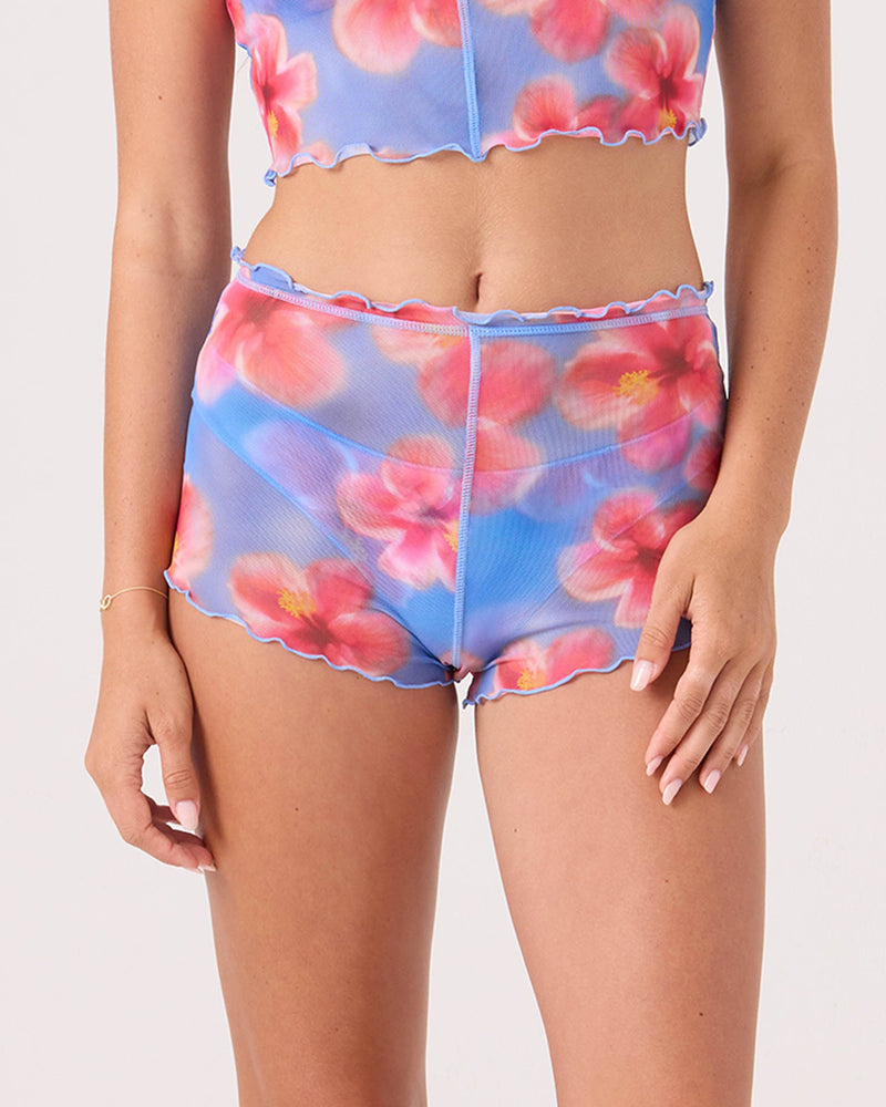 A person models the Roxy Hibiscus Daze Mesh Shortie Cheeky Bikini Bottom in Blue Haze Hibiscus, featuring ruffled hems and a matching sheer crop top, against a plain light background.