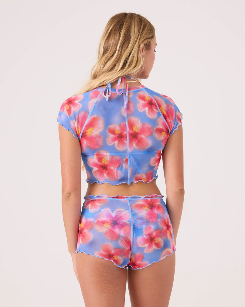A woman with long blonde hair faces away, wearing Roxy's Hibiscus Daze Mesh Shortie Cheeky Bikini Bottom featuring vibrant pink and red hibiscus flowers. Her top is sheer blue, and the plain background is light-colored.