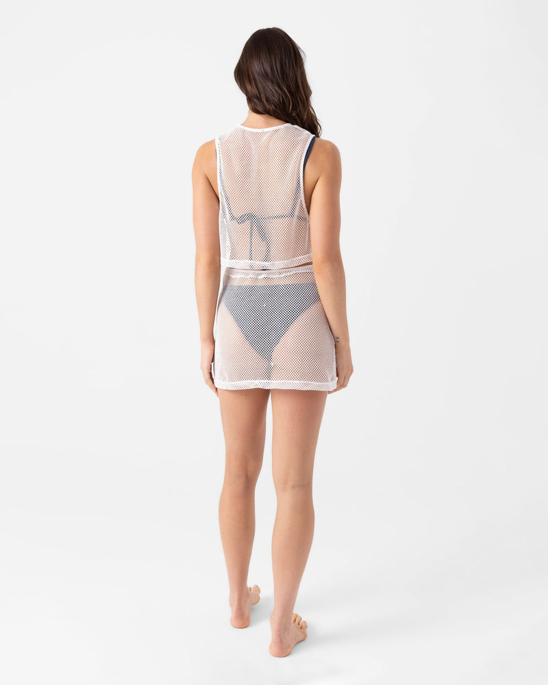 A woman, barefoot and facing away against a white backdrop, models the Roxy Don't Mesh With Me Skirt Cover Up—a sheer white mesh skirt layered over a dark bikini for a beach-ready look.