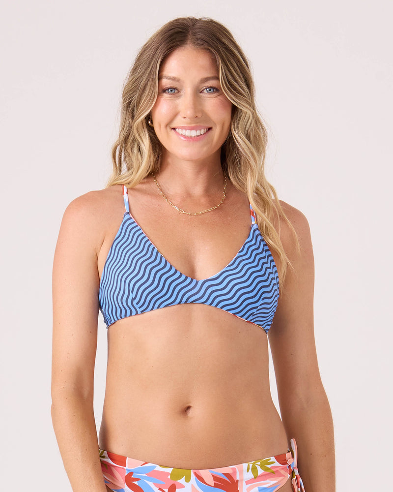 A woman smiles, wearing the Roxy Ripple Effect Reversible Athletic Triangle Bikini Top in Blue Multi with a criss cross back and floral bottoms, posing against a plain light background.