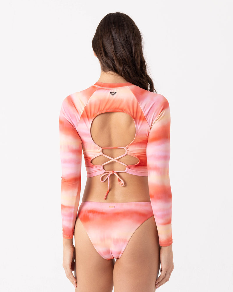 A woman with long brown hair models the Roxy Palm Before The Storm Crop Rashguard, a pink and orange tie-dye long sleeve bikini set with an open back and crisscross lace-up details in recycled nylon, against a white background.