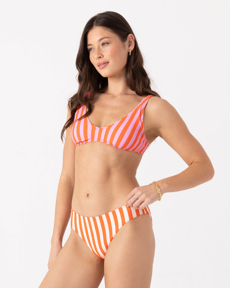 A woman models the Ya Stripe Me Crazy Reversible Scoop Bralette Bikini Top by Roxy in pink and orange stripes, standing against a plain white background and looking to the side with a relaxed expression.