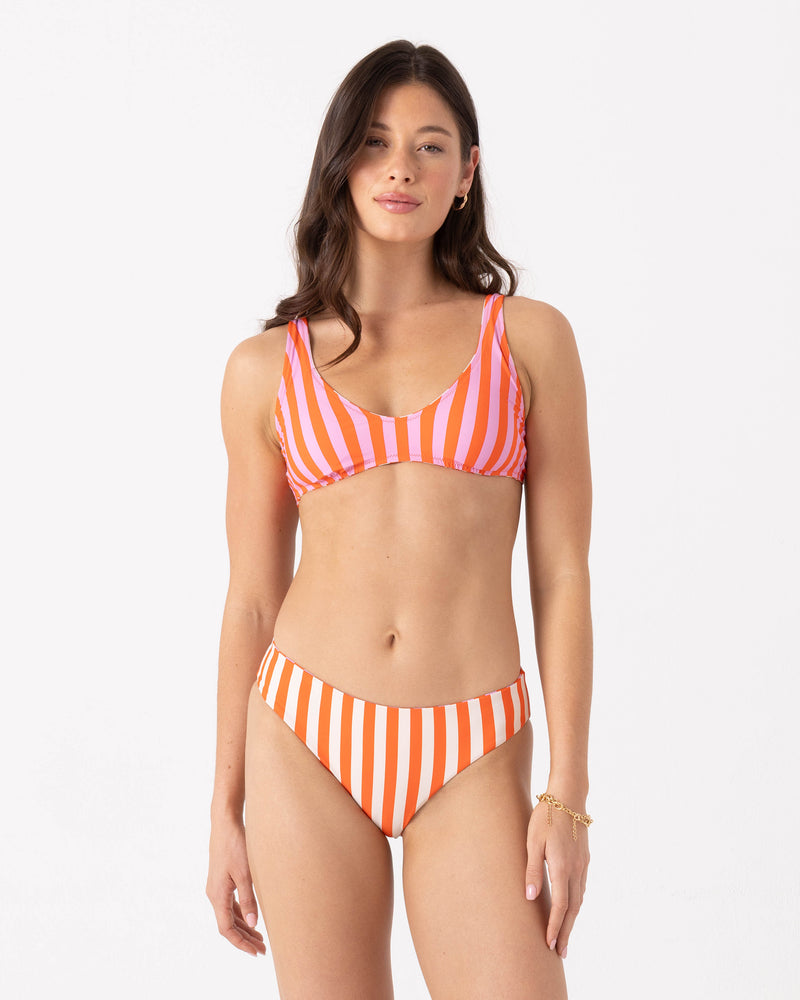 A woman models the Roxy Ya Stripe Me Crazy Reversible Scoop Bralette Bikini Top, featuring pink and orange stripes and recycled fabric, against a plain white background with a relaxed expression.