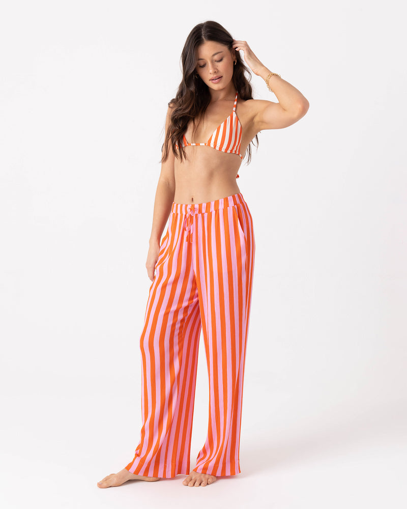 A woman wears a matching bikini top and Roxy Ya Stripe Me Crazy Beach Pant with a drawcord elastic waist, both featuring vertical orange and pink stripes.