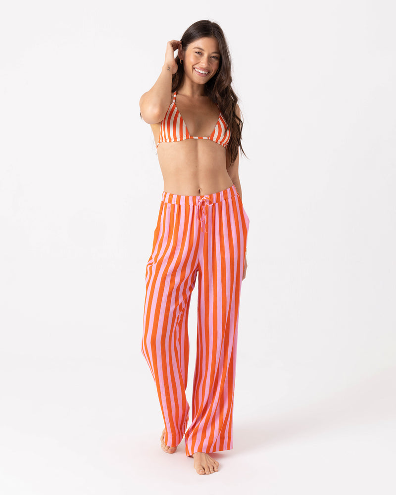 A woman wears a matching bikini top and Roxy Ya Stripe Me Crazy Beach Pants, which feature a vibrant orange and pink striped wide-leg design and a drawcord elastic waist.