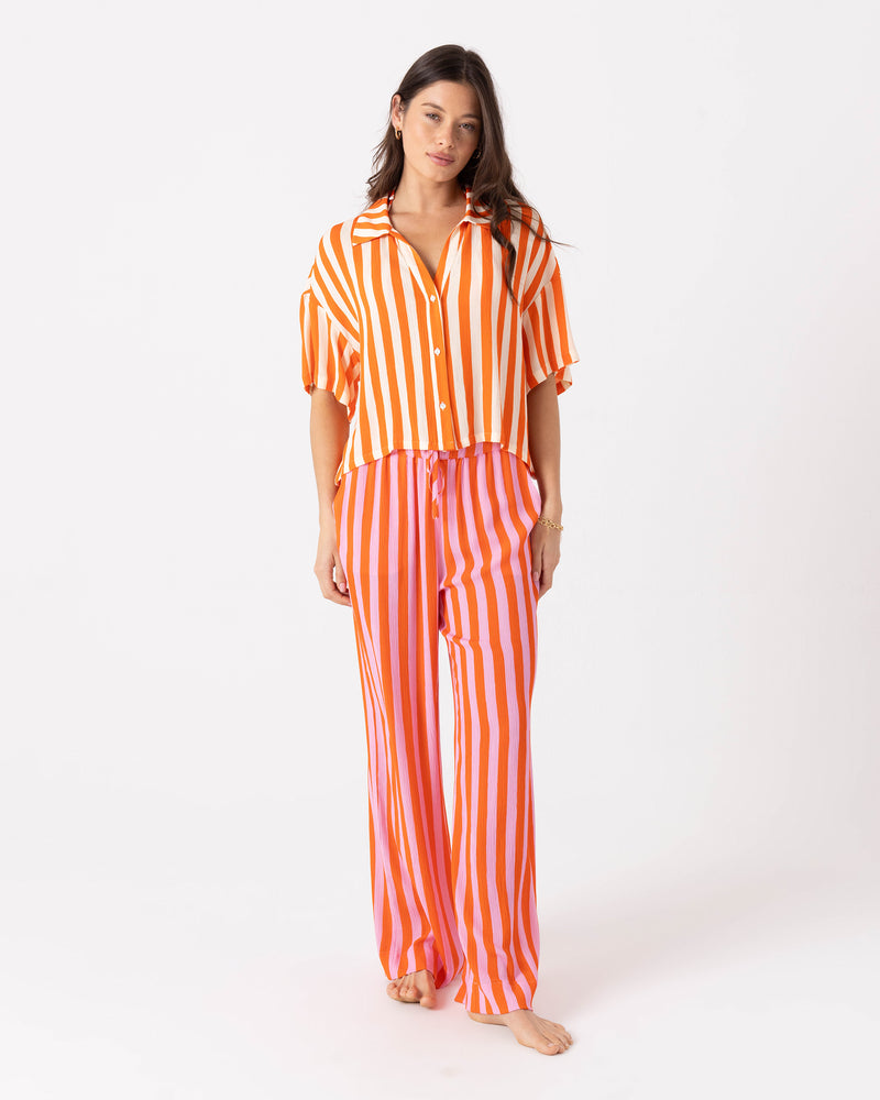 A person wears an orange and white vertically striped button-down shirt paired with matching pink and orange Ya Stripe Me Crazy Beach Pant featuring a drawcord elastic waist.