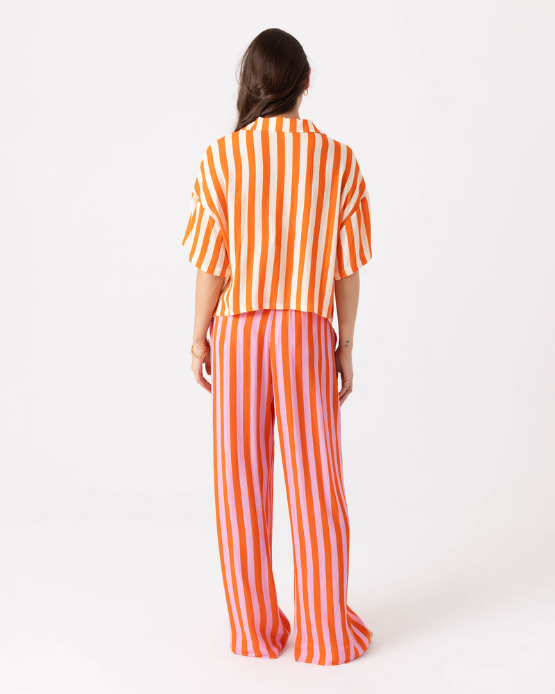 A person seen from the back wearing a short-sleeved collared shirt with orange and white vertical stripes and Roxy Ya Stripe Me Crazy Beach Pant with an elastic waist in orange and pink vertical stripes.