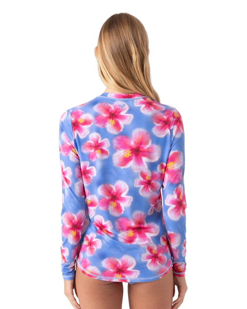 A woman with long blonde hair is seen from the back, wearing the Roxy Hibiscus Daze Sea Skippin Rashguard featuring a blue background and pink hibiscus print, standing against a plain white backdrop.