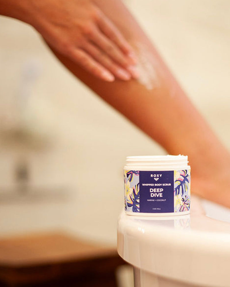 A ROXY Deep Dive Body Butter & Scrub Bundle rests on the bathtub edge as someone exfoliates their leg in the background, enjoying marine notes and deeply hydrated skin.