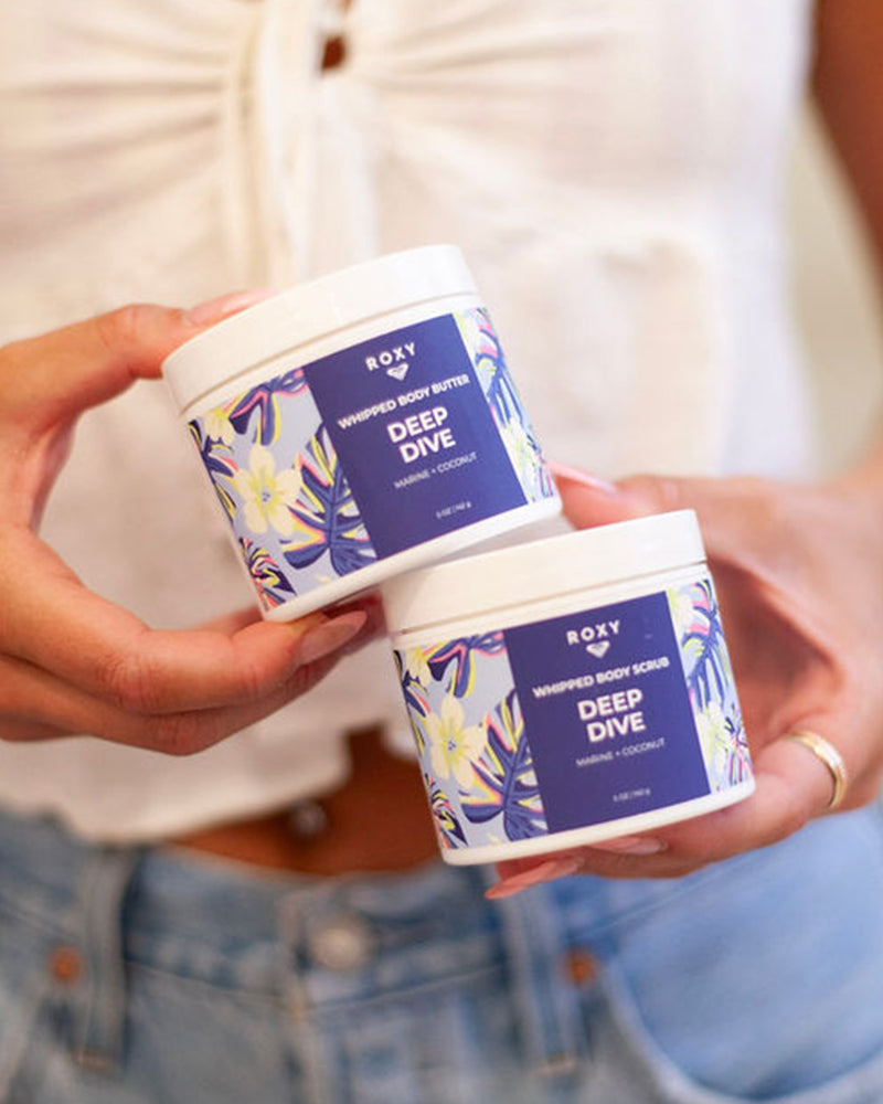 A person in a white top and blue jeans holds the ROXY Deep Dive Body Butter & Scrub Bundle with blue floral labels, perfect for exfoliating and hydrating the skin.