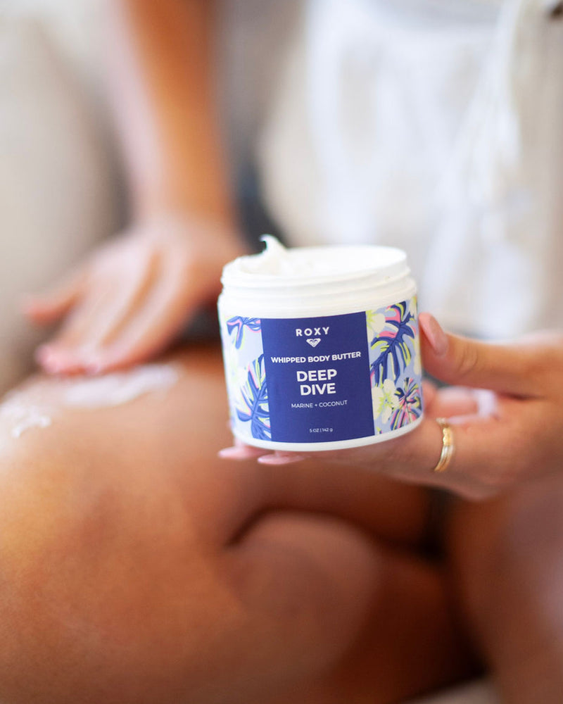 A person applies ROXY’s Deep Dive Body Butter & Scrub Bundle to their leg, holding a jar with a colorful floral design, leaving skin soft, hydrated, and infused with fresh marine notes.