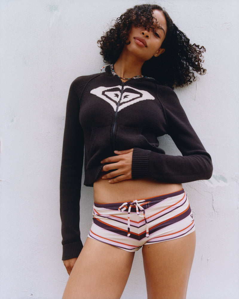 A woman with curly hair poses against a white wall, wearing a black zip-up hoodie with a white logo and the ROXY x Madhappy Swim Short Cheeky Bikini Bottom in Multi by Roxy x Madhappy, featuring a drawstring and sporty stripes.