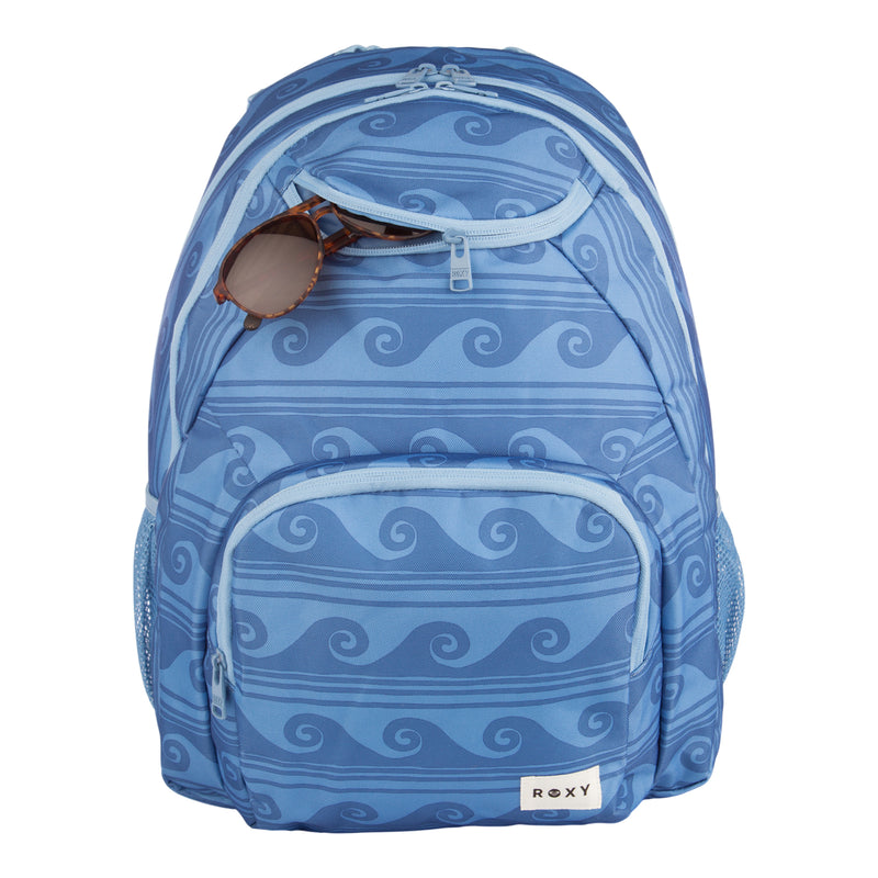 The ROXY Shadow Swell Backpack features a blue wave pattern, front zipper pockets, mesh side pockets, and brown sunglasses partially tucked into the top compartment. A small “ROXY” label appears on the lower front pocket.