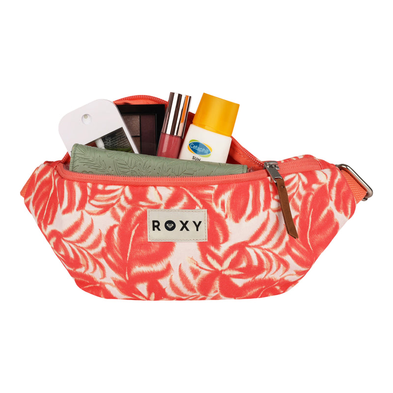A pink and cream patterned ROXY Vanilla Smoothie Crossbody Bag unzipped, revealing makeup items, a sunscreen bottle, a green case, and a white compact inside—perfect for slipping into your favorite cotton crossbody with an adjustable strap for easy, hands-free carrying.