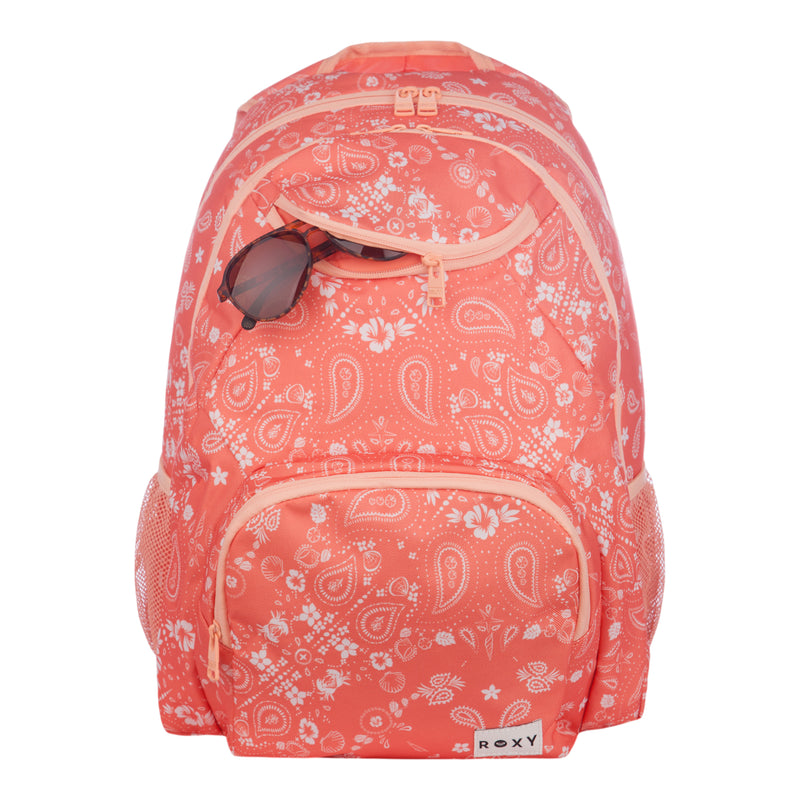 The Roxy Shadow Swell Backpack features a coral color with white paisley patterns, multiple zippered compartments, and a pair of sunglasses tucked into the top pocket. The front displays the Roxy label.