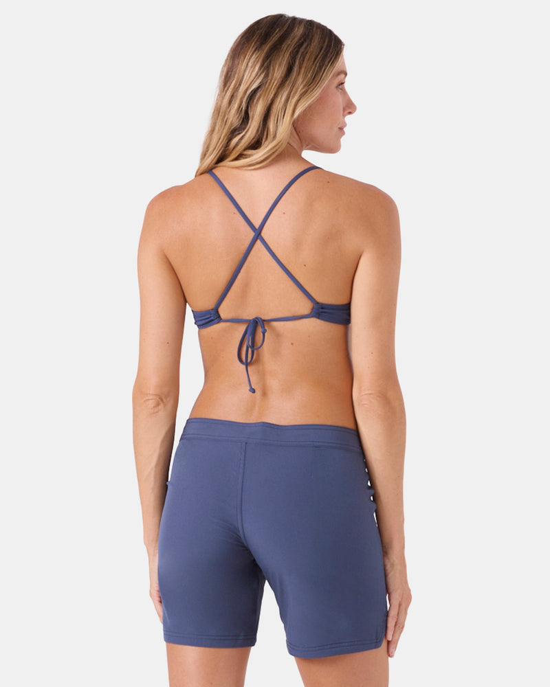 A woman faces away, modeling Roxy’s To Dye 7" Boardshorts and a blue crisscross tie-back bikini top. Her light brown hair is down, set against a plain white background.