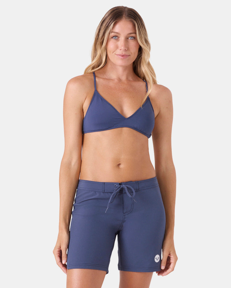 A woman with long, wavy blonde hair models Roxy's To Dye 7" Boardshort and a matching blue bikini top, standing and facing forward against a plain white background.