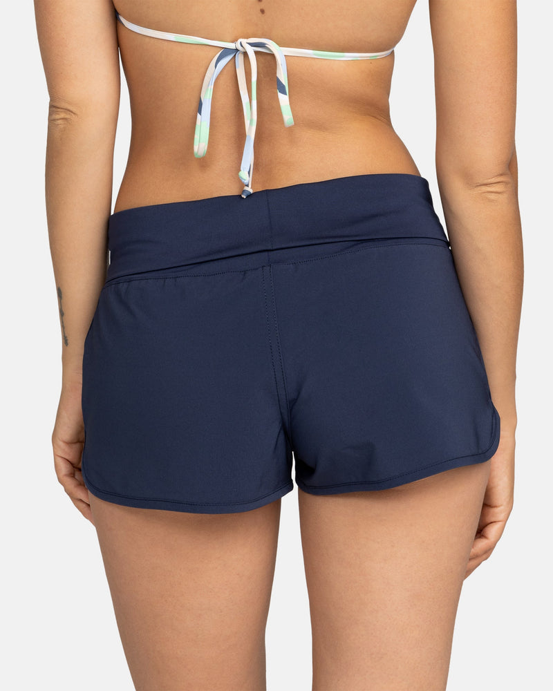 A person is shown from the back in a bikini top and Roxy Endless Summer Boardshorts in Mood Indigo. The background is plain and light-colored.