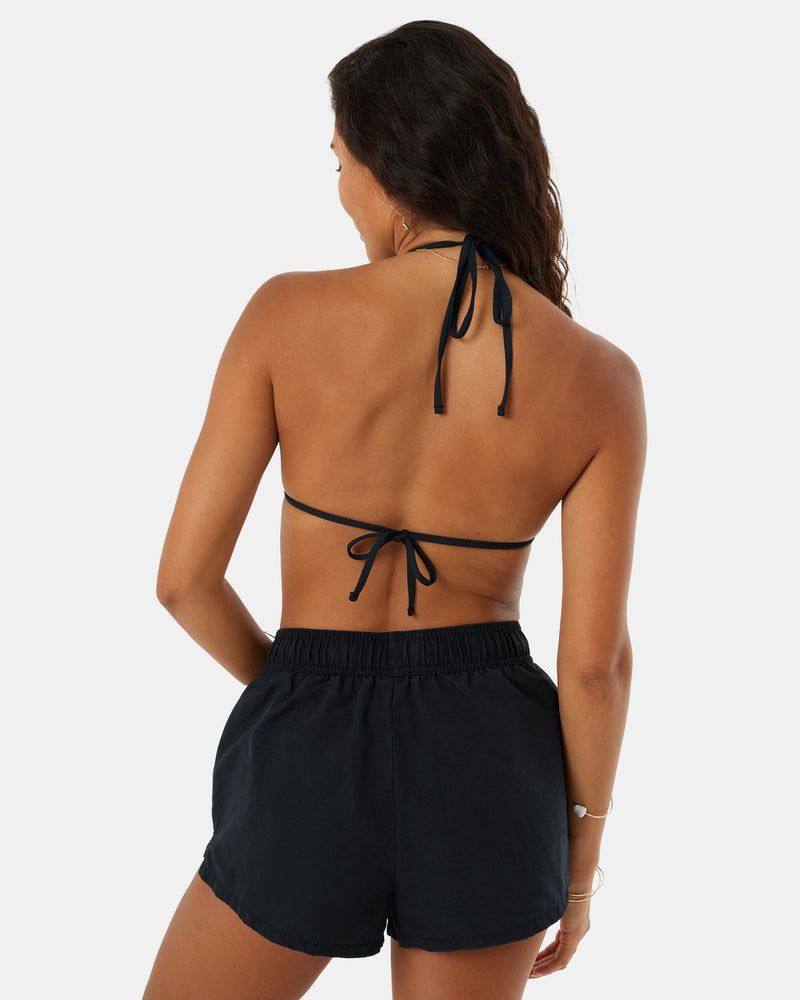 A woman with long brown hair is shown from the back, wearing the Roxy No Bad Waves black halter bikini top and 4-way stretch cover-up boardshorts, with one hand on her hip against a plain white background.