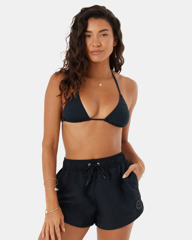 A woman with long wavy hair wears the Roxy No Bad Waves black bikini top and 4-way stretch cover-up boardshorts, standing against a plain light background with one hand in her pocket, gazing confidently at the camera.