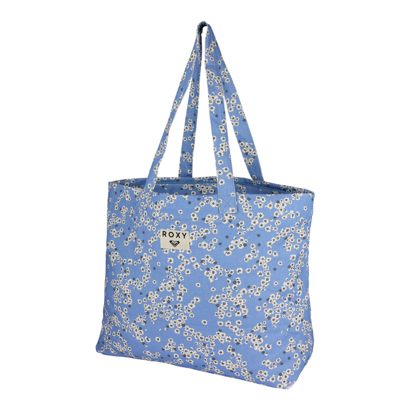 The ROXY Sweeter Than Honey Tote Bag showcases a delightful white daisy print, two durable shoulder straps, and a sewn-on ROXY brand label on the front. Crafted from recycled cotton for an eco-friendly touch, this tote bag stands upright against a clean white background.