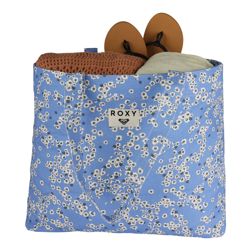A blue ROXY Sweeter Than Honey Tote Bag with a white floral pattern, filled with a brown knitted item, tan sandals, and a light green rolled towel. This eco-friendly ROXY tote bag is perfect for carrying your essentials in sustainable style.