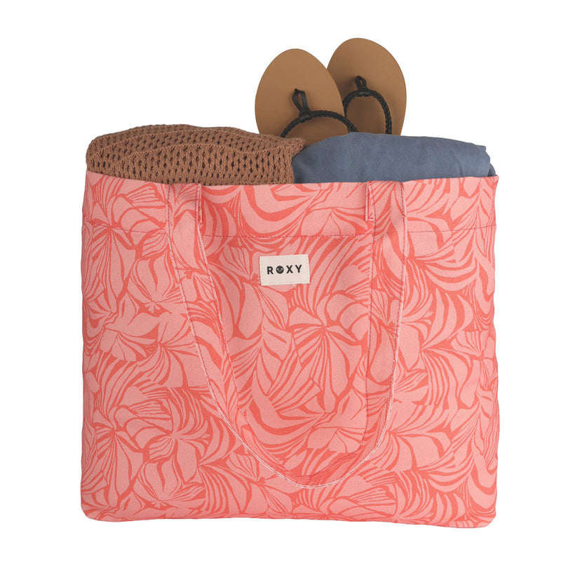 A pink floral Sweeter Than Honey Tote Bag by ROXY, made from eco-friendly recycled cotton, holds a brown knit item, tan sandals, and a folded blue garment—each visible through the bag’s open top.