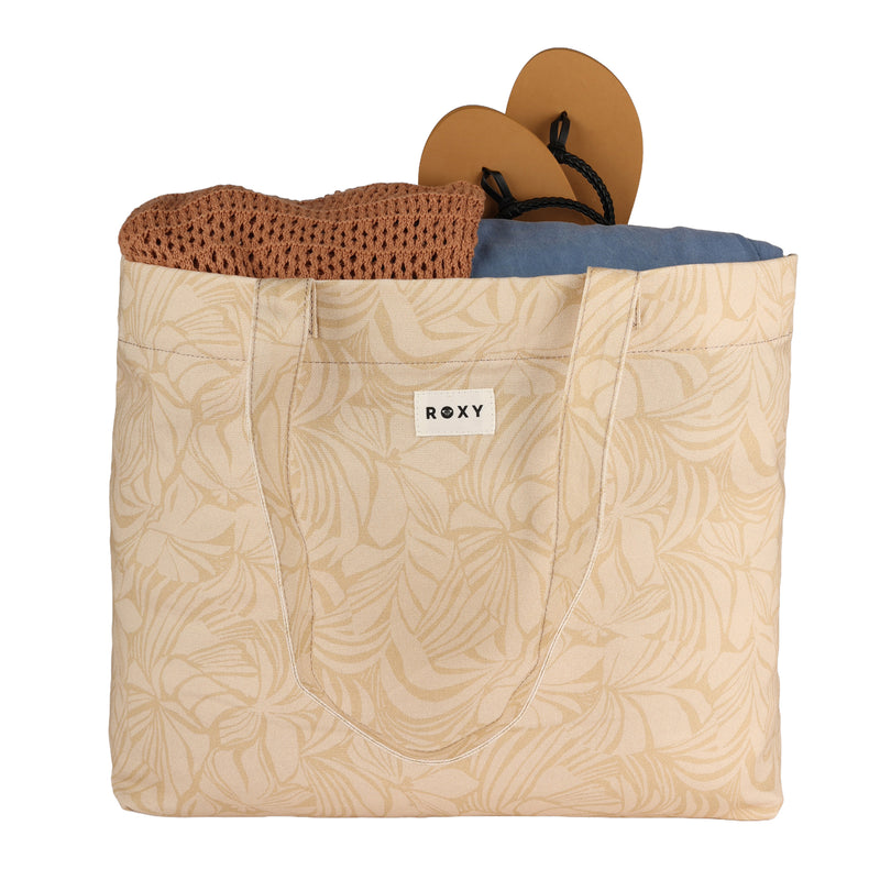 A beige patterned ROXY Sweeter Than Honey Tote Bag, crafted from recycled cotton, carries a brown knit sweater, a blue cloth, and a pair of tan flip-flops with black straps. This eco-friendly tote from ROXY is perfect for stylishly transporting your everyday essentials.