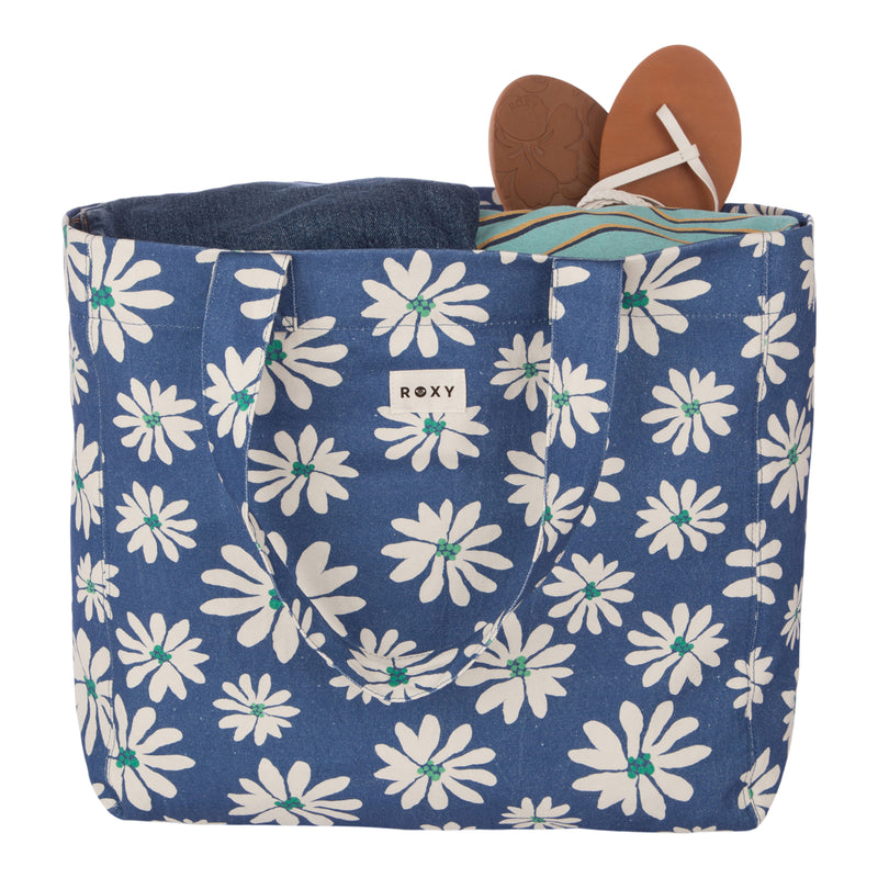 Introducing the Roxy Anti Bad Vibes Tote Bag—a chic blue accessory adorned with white daisy prints and featuring dual handles along with a signature Roxy label on the front. Crafted from recycled cotton, this stylish tote comes pre-packed with a pair of tan sandals and folded blue jeans.