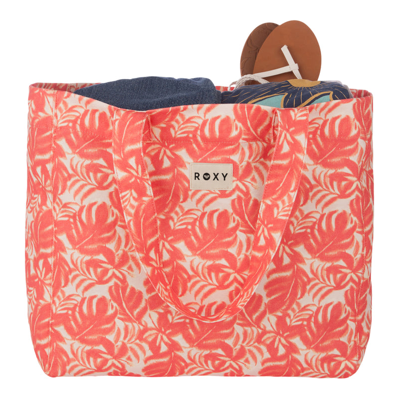 A front-facing view of the ROXY Anti Bad Vibes Tote Bag in a pink and white tropical pattern, stylishly filled with clothing and a pair of brown sandals featuring white straps.